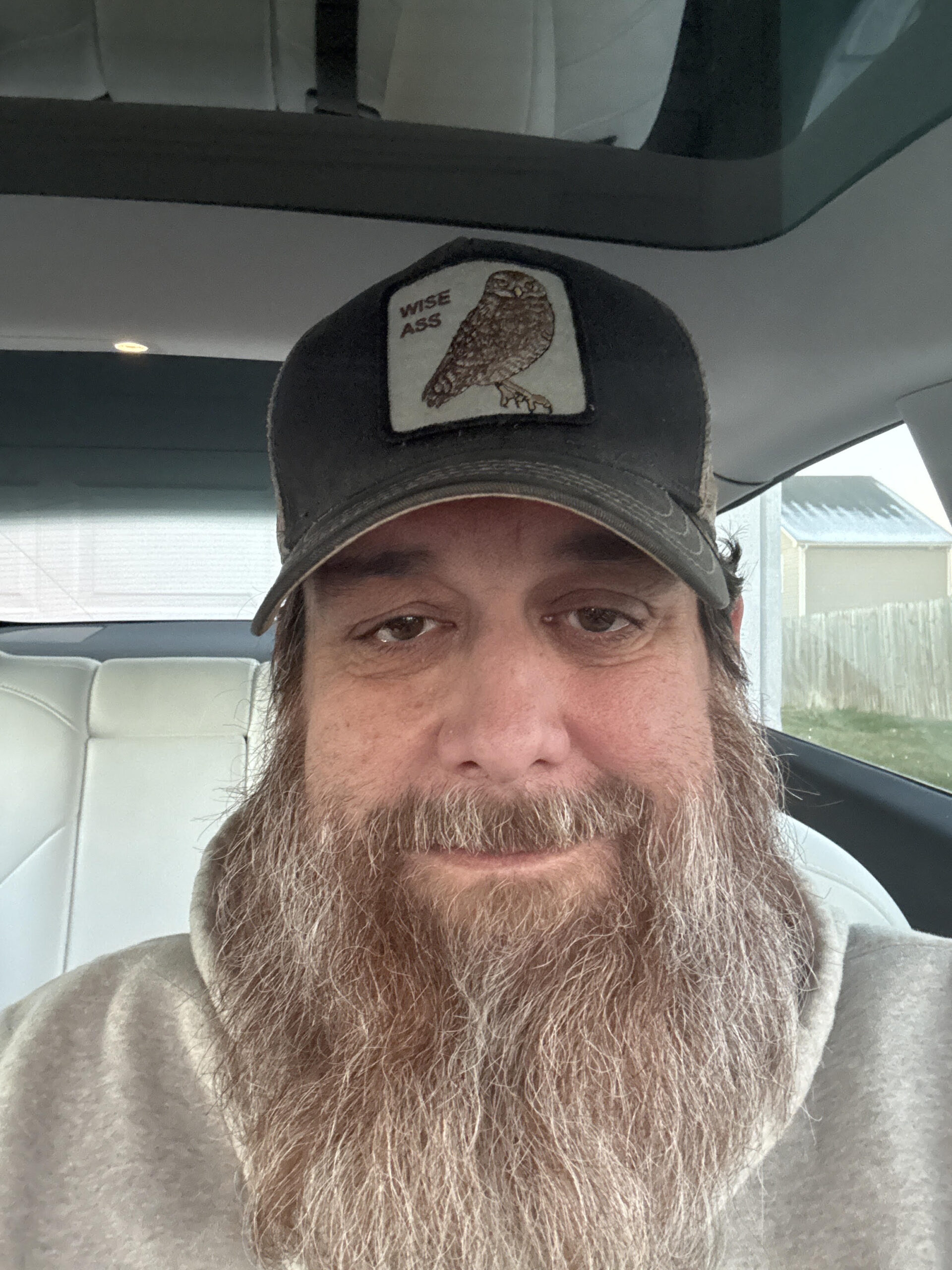 Portrait of Todd, a bearded U.S. militaryveteran wearing a hat with a “Wise Ass” owl patch, seated in a vehicle.